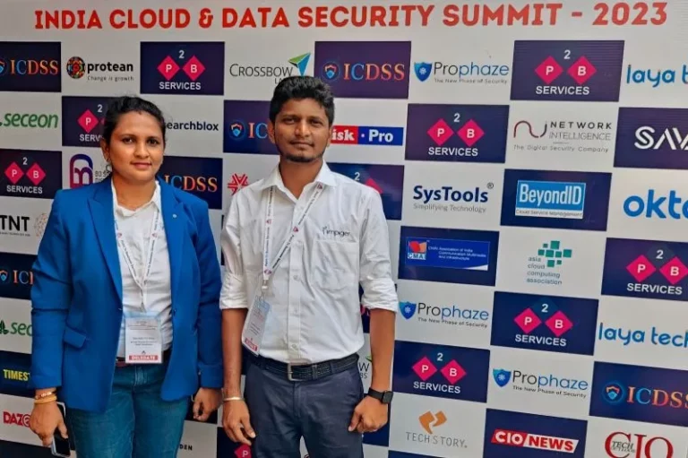 Impiger team members wearing professional attire, standing in front of a conference backdrop with the text 'India cloud and Data Security Summit 2023'. They are engaged in conversation and indicating their participation in the event.